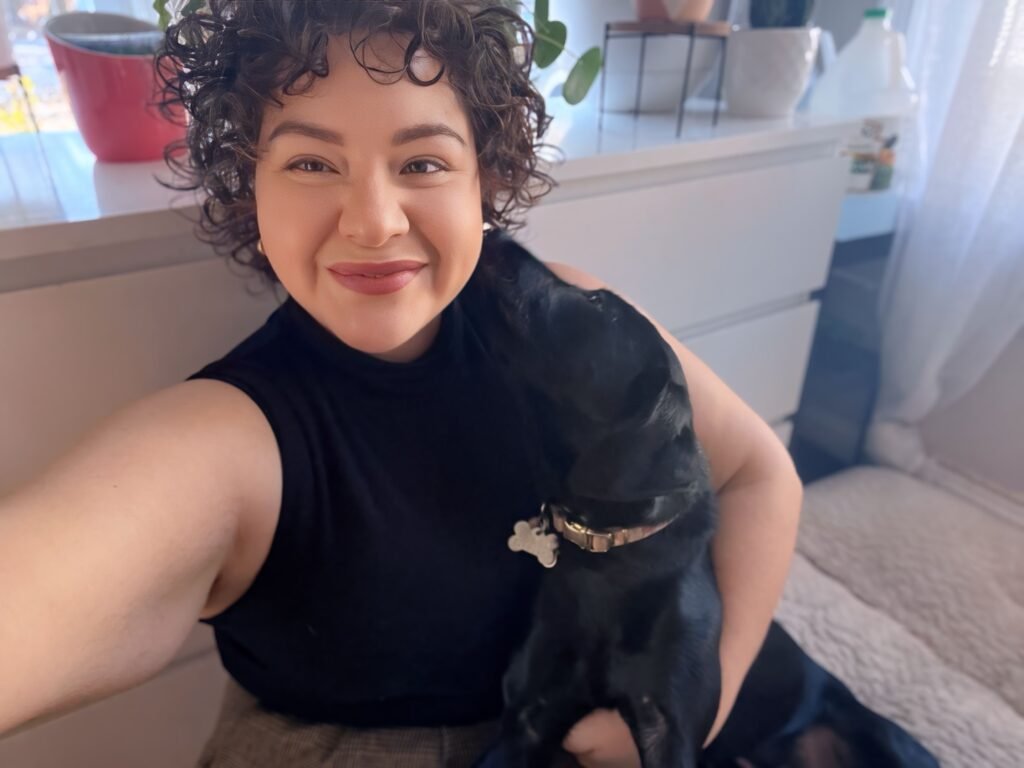 Smiling woman with short curly hair holding a black dachshund indoors near a window with natural light.